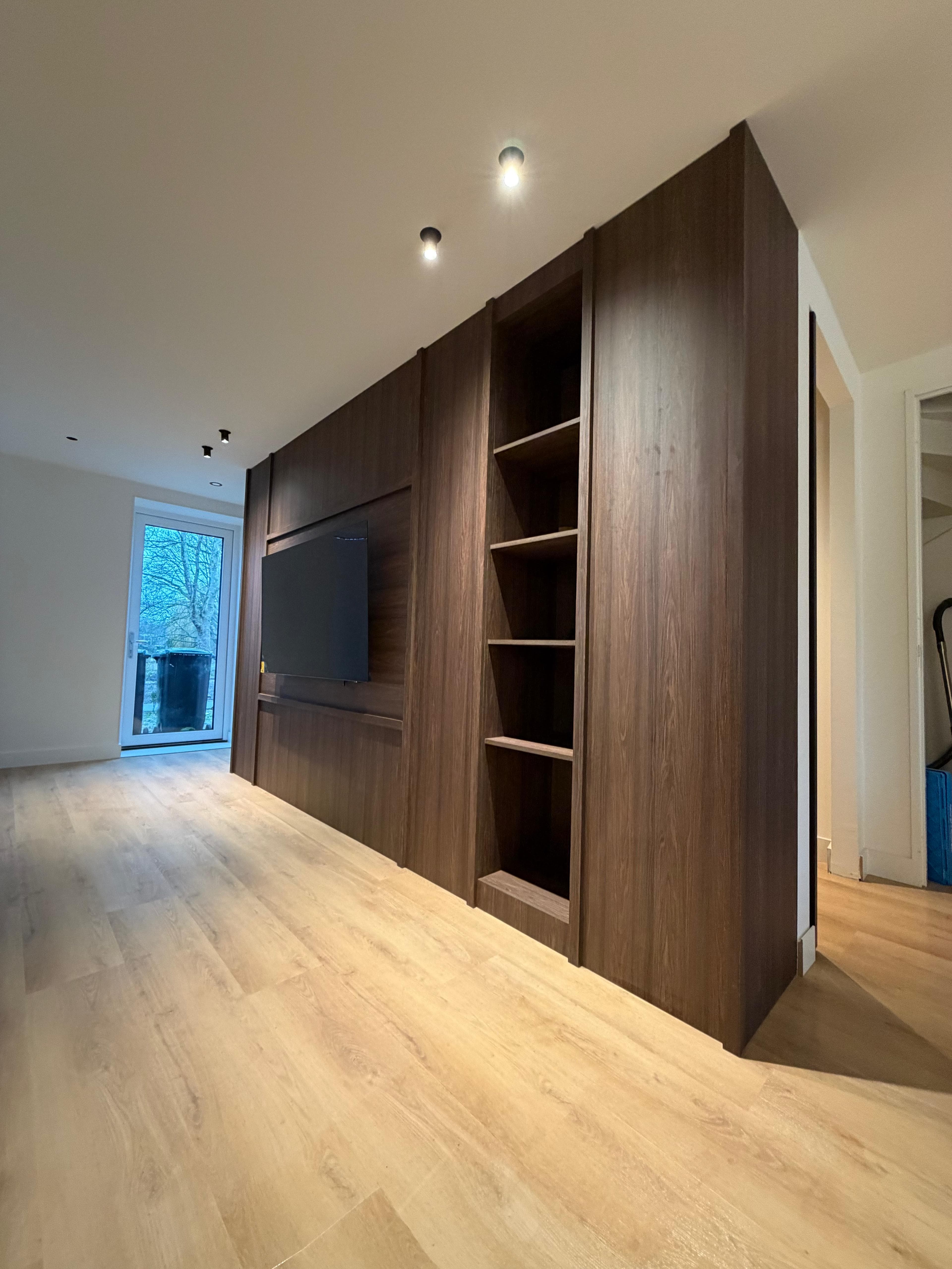 Opgeleverde inbouwkast van donker hout, van vloer tot plafond, met open vakken en een ingebouwde tv-wand.